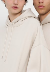 Gros plan de deux personnes portant des sweat-shirts à capuche beige clair avec cordons, montrant partiellement les visages et les épaules sur un fond uni.