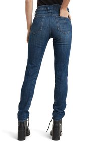 Kvinna i mörkblå skinny jeans och svarta klackstövlar, med en hand i bakfickan, visad från baksidan på vit bakgrund.