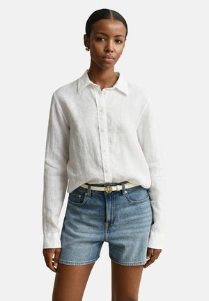 Frau trägt ein weißes, langärmliges Hemd mit Knöpfen, das in hoch taillierte blaue Jeansshorts mit weißem Gürtel und goldener Schnalle gesteckt ist.