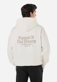 Sudadera crema clara con una gran impresión en la parte posterior en texto beige oscuro. Cuenta con capucha con cordón y puños acanalados. Corte casual y oversize.
