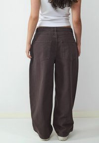 Szerokie brązowe dżinsy typu wide-leg z wysokim stanem, z dwiema tylnymi kieszeniami i gładką fakturą. Noszone z dopasowanym białym topem bez rękawów.