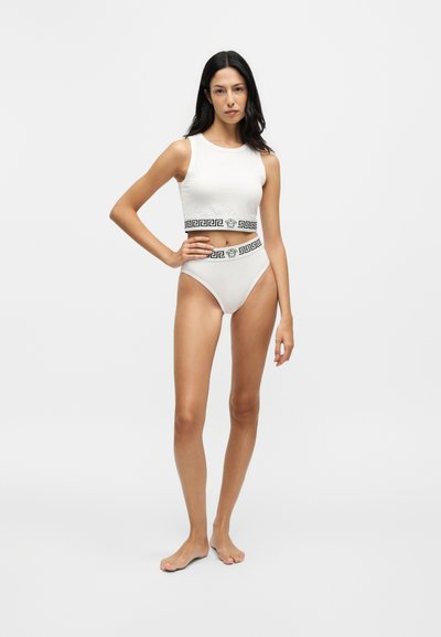 Femme aux longs cheveux foncés, debout pieds nus, portant un crop top blanc sans manches et un sous-vêtement taille haute avec des bandes décoratives noires à la taille.