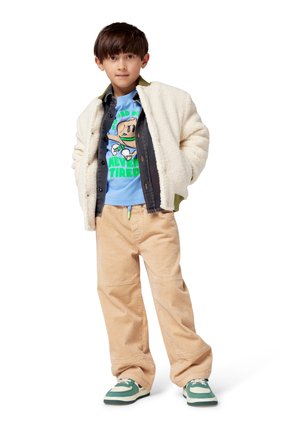 Child wearing a cream fleece jacket over a blue graphic t-shirt, beige corduroy pants, and green-accented sneakers.