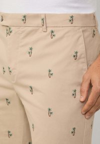 Beige trousers with embroidered small green palm trees, featuring a smooth texture and classic design with a button closure at the waist.