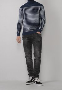 Maglione a collo alto a righe navy con strisce bianche, abbinato a jeans scuri slim-fit e sneaker nere con dettagli bianchi.