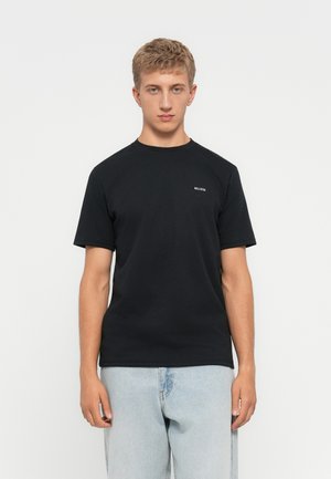 Ung mann iført svart Hollister t-skjorte og lyseblå jeans, står mot en ensfarget hvit bakgrunn.