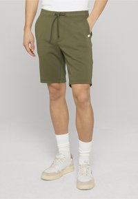 Shorts en coton vert olive avec une taille élastique et un cordon de serrage. Comprend des poches latérales et une étiquette logo. Texture lisse, design arrivant au genou.
