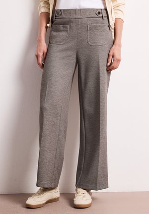 Vrouw die grijze broek met wijde pijpen en voorzakken draagt, samen met crèmekleurige sneakers, staand tegen een witte muur.