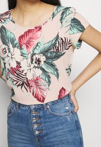 Haut à manches courtes imprimé floral, en tissu rose pâle avec des feuilles vertes et rouges, associé à un jean en denim bleu taille haute avec des boutons.
