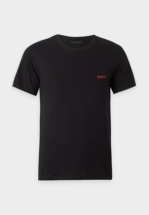 Mann steht barfuß, trägt ein schwarzes T-Shirt und Jogginghosen mit kleinen roten Hugo-Logos, die Hände in den Taschen, vor schlicht weißem Hintergrund.