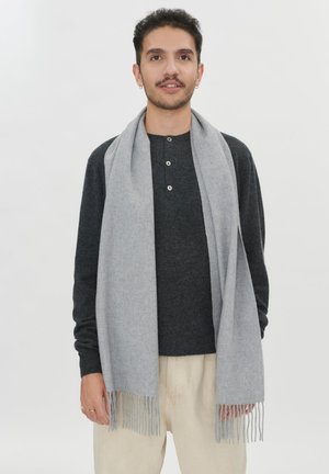 Mann trägt dunkelgraues Langarmshirt, hellgrauen Schal mit Fransen und beige Hose, steht vor einem schlichten weißen Hintergrund.