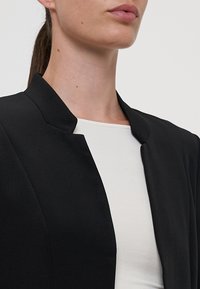 Fekete zakó magas gallérral, sima anyagból készült. Egy fehér felsőre van felvéve, amely karcsú sziluettet és minimalista dizájnt mutat.