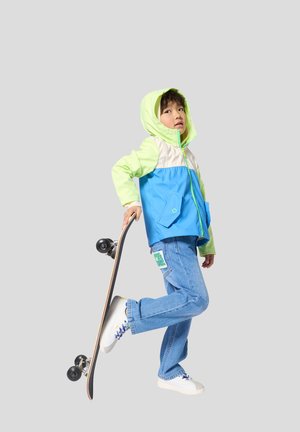 Kleurrijke windjack met groene en blauwe panelen, voorzien van een capuchon en een voorzak. Lichtblauwe spijkerbroek met een merkpatch, witte sneakers.