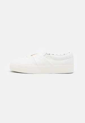Baskets blanches à enfiler avec une tige en toile texturée, présentant un design noué à l'avant et une semelle en caoutchouc.
