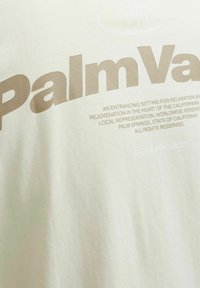 Svijetlosmeđa majica kratkih rukava s velikim smešnim slovima koja kaže "Palm Valley" i deskriptivnim tekstom ispod u manjem fontu. Mekani pamučni materijal.