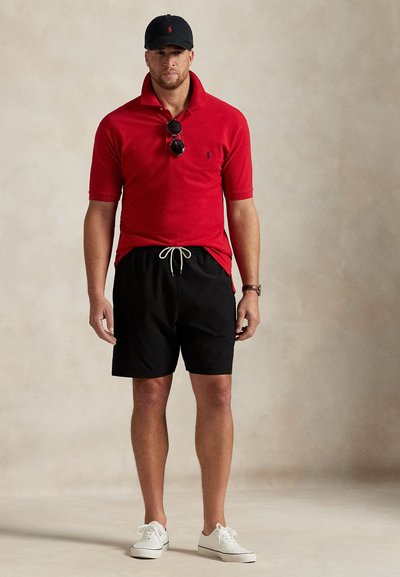 Raudonas polo marškinėlis su apykakle, juodi šortai su raišteliu, balti sportbačiai ir juoda kepurė su logotipu; ant marškinėlio padėtos saulės akiniai.