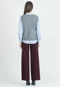 Grijze gebreide vest over een blauw gestreept shirt, gecombineerd met ruime maroon broek en zwarte hakken, gezien vanuit de rug.