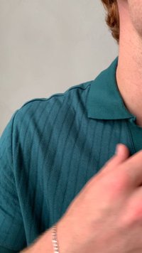 Mėlynas tekstūruotas polo marškinėlis su vertikaliomis stulpeliais ir klasikinėmis apykaklėmis, iš dalies matomas su ranka, liečiančia apykaklės sritį.
