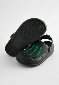 Sabots noirs avec des accents en forme d'os verts sur le dessus. Fabriqués en matériau flexible, dotés d'une semelle texturée pour une meilleure adhérence et soutien du pied.