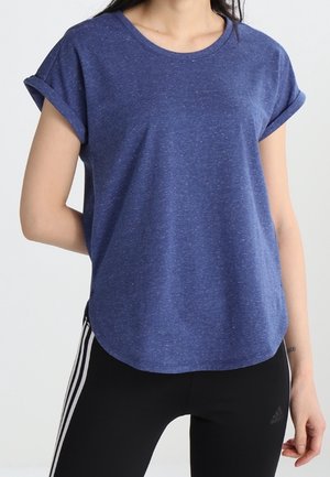 Kobieta w niebieskim T-shircie z podwiniętymi rękawami i czarnych legginsach Adidas z białymi paskami, stojąca na jednolitym tle.