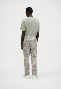 Olive zöld pamut póló, világosszürke terepmintás cargo nadrággal párosítva, amely több zsebbel és elasztikus derékrésszel rendelkezik. A megjelenést fehér sneakers egészítik ki.