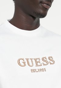 T-shirt bianca con logo "GUESS" ricamato marrone e dettaglio "EST. 1981" sul davanti, con collo a giroclassico.