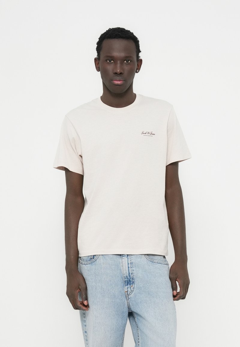 Ανοιχτό μπεζ μπλουζάκι με στρογγυλή λαιμόκοψη, κοντά μανίκια και διακριτικό λογότυπο στο στήθος. Συνδυασμένο με ανοιχτό μπλε τζιν παντελόνι.