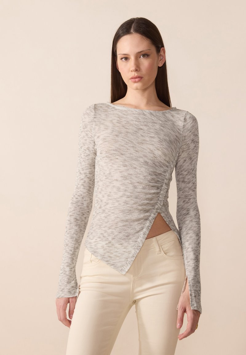 Top lung cu mâneci lungi de culoare gri deschis, cu un model texturat, având un tiv asimetric și plissare pe o parte, asociat cu pantaloni de culoare deschisă.