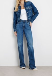 Veste en denim bleu moyen avec des fermetures à boutons, deux poches poitrine et des manches longues. Assortie à un jean taille haute à jambes larges dans une teinte similaire.