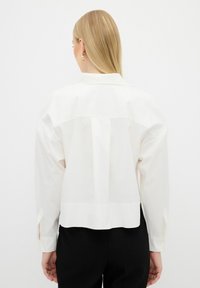 White structured shirt with a collar, long sleeves, and button cuffs. Features a yoke detail on the back and a slightly cropped hem.