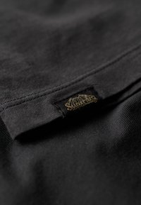 Tela negra con una textura suave, con una etiqueta cosida que presenta un diseño de concha bordado en dorado en el dobladillo.
