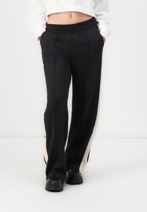 Pantalons de sport noirs avec un design à jambes larges, dotés d'une bande latérale blanche et d'une taille élastique, fabriqués en un matériau lisse et texturé.