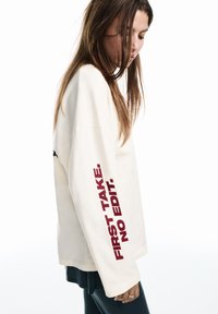 Crèmekleurige sweatshirt met lange mouwen en bordeauxrode tekst "FIRST TAKE. NO EDIT."; heeft een ontspannen pasvorm en een zachte textuur.