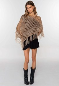 Poncho de punto abierto en color beige con detalle de flecos en el dobladillo. Estilizado sobre una falda negra y combinado con botas de cowboy negras.