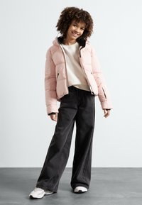 Padded rosa Jacke mit Kapuze, schwarzer Innenseite, Front-Reißverschluss und Seitentaschen. Kombiniert mit weiten schwarzen Hosen und weißen Sneakers.