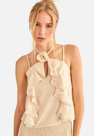 Mujer rubia con top beige sin mangas con bordes de encaje con volantes y detalle floral en el cuello, combinada con falda beige texturizada.