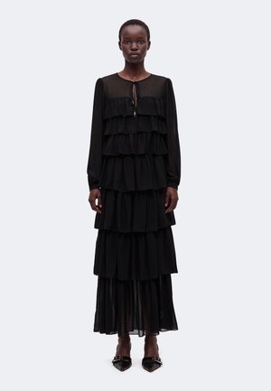 Femme portant une longue robe noire à volants avec manches longues et un lien au niveau du col, accompagnée de chaussures noires à bout pointu.