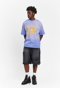 Løs passform lavendel tie-dye t-skjorte med gul tekst og grafikk, paret med mørke cargo-shorts og svarte snøringstøvler.