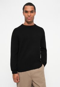 Sweater de malha de manga longa preto com um decote redondo. Textura suave e corte descontraído. Combinado com calças de tom claro.