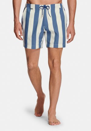 Man die blauw-witte zwemshorts met verticale strepen en een vooraanliggende trekkoord draagt, lopend op blote voeten op een witte achtergrond.