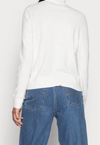 Pull en maille blanche à col roulé avec manches longues, avec un ourlet côtelé. Associé à un jean en denim bleu taille haute, présentant des poches arrière en forme de cœur.