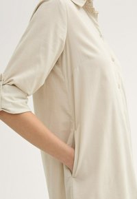 Camicia button-up beige chiaro con colletto, maniche arrotolate, tasche laterali e una texture morbida e liscia.