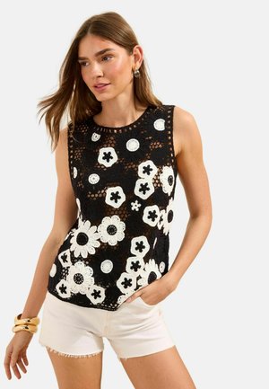 Femme portant un top en crochet noir sans manches avec des motifs floraux blancs, un short blanc, des bracelets dorés et des boucles d'oreilles, posant avec une main dans la poche.