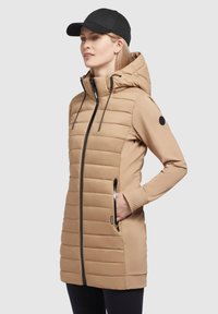 Beige krēpju jaka ar rāvējslēdzēju priekšpusē, kapuci un ribotas aproces. Iezīmē melnas rāvējslēdzēju detaļas un nozīmīti uz piedurknes. Apvienota ar melnu cepuri.