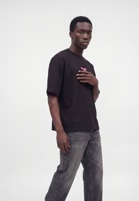 Jeune homme debout avec une main sur la poitrine, portant un t-shirt noir avec un texte rose et un jean gris délavé sur un fond blanc uni.