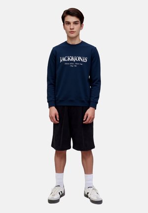 Navy blue crewneck sweatshirt with white "JACK&JONES" text, paired with black shorts and white sneakers, featuring white socks.