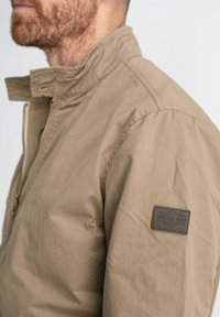 Beige zippered jacket with a stand collar, made of smooth, lightweight fabric. Features a stitched logo patch on the left sleeve.