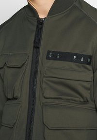 Olivgrön bombarjacka med dragkedja framtill, räfflad krage och fyra fickor fram. Har en svart tygpatch med text. Slät textur.