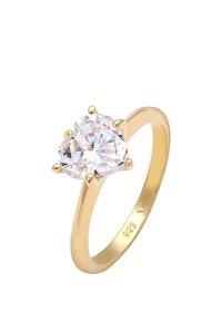 Anello placcato oro con un grande pietra trasparente a forma di cuore, fissata con quattro griffe; la fascia è liscia con una finitura lucida.