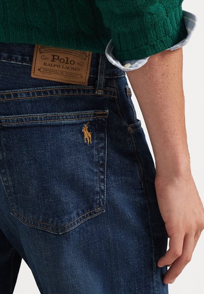 Dark blue denim jeans with a tan leather label featuring "Polo Ralph Lauren." Gold embroidery of a horse on the rear pocket. Textured fabric.